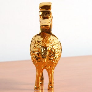 May include: A gold-colored metal figurine of a ram with a detailed, textured surface. The ram is standing on a light brown surface.