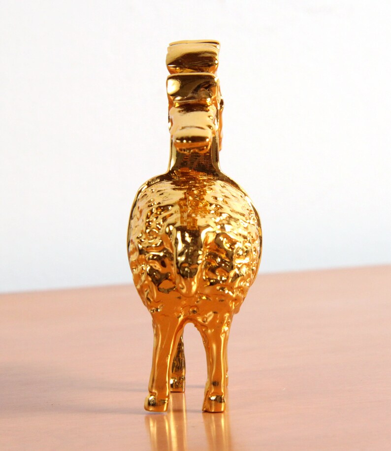 May include: A gold-colored metal figurine of a ram with a detailed, textured surface. The ram is standing on a light brown surface.