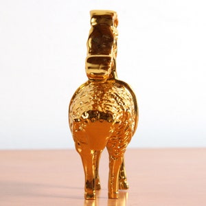 May include: A gold-colored metal figurine of a sheep with a curly fleece. The sheep is standing on a wooden surface.