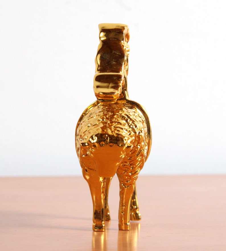 May include: A gold-colored metal figurine of a sheep with a curly fleece. The sheep is standing on a wooden surface.
