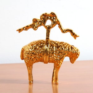 May include: A gold-colored metal sculpture of a sheep with a stylized tree branch on its back. The sheep is standing on a wooden surface.