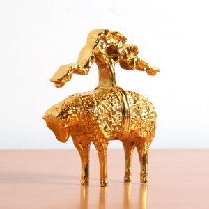 May include: A gold-colored metal sculpture of a sheep with a stylized tree growing from its back. The sculpture has a textured surface and is detailed with intricate carvings.