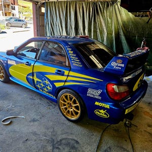 K&ouml;nnte beinhalten: Ein blaues Subaru Impreza WRX STI Sportwagen mit gelben und blauen Grafiken. Das Auto hat goldene Felgen, einen Heckspoiler mit "Corona Extra"-Text und verschiedene Sponsorenaufkleber. Das Auto steht in einer Garage.