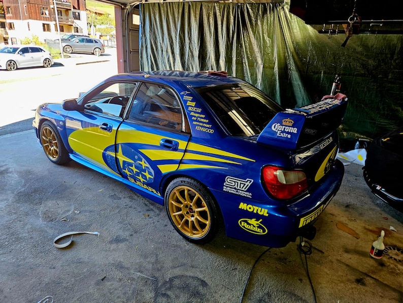 K&ouml;nnte beinhalten: Ein blaues Subaru Impreza WRX STI Sportwagen mit gelben und blauen Grafiken. Das Auto hat goldene Felgen, einen Heckspoiler mit "Corona Extra"-Text und verschiedene Sponsorenaufkleber. Das Auto steht in einer Garage.