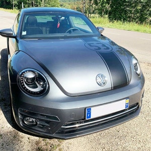 Puede incluir: Un Volkswagen Beetle gris con una franja de carreras negra y el número "53" en el capó. El coche tiene una matrícula europea con las letras "F" y "71".