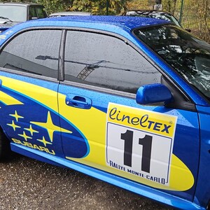 K&ouml;nnte beinhalten: Blauer Subaru Rallye-Wagen mit gelben und blauen Grafiken, goldenen R&auml;dern und einem Heckspoiler. Das Auto hat die Nummer 11 auf der Seite, zusammen mit dem Text "linelTEX" und "Rallye Monte Carlo."
