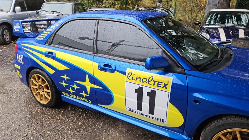 K&ouml;nnte beinhalten: Blauer Subaru Rallye-Wagen mit gelben und blauen Grafiken, goldenen R&auml;dern und einem Heckspoiler. Das Auto hat die Nummer 11 auf der Seite, zusammen mit dem Text "linelTEX" und "Rallye Monte Carlo."