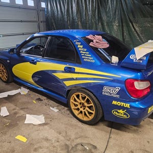 K&ouml;nnte beinhalten: Ein blaues Sportwagen mit gelben und wei&szlig;en Akzenten, goldenen Felgen und verschiedenen Sponsorenaufklebern. Das Auto hat einen Heckspoiler mit "Corona Extra"-Branding. Das Auto steht in einer Garage.