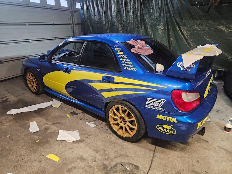 K&ouml;nnte beinhalten: Ein blaues Sportwagen mit gelben und wei&szlig;en Akzenten, goldenen Felgen und verschiedenen Sponsorenaufklebern. Das Auto hat einen Heckspoiler mit "Corona Extra"-Branding. Das Auto steht in einer Garage.