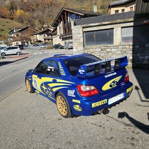 K&ouml;nnte beinhalten: Ein blaues Subaru Rallye-Auto mit gelben Akzenten und goldenen R&auml;dern steht auf einer Stra&szlig;e. Das Auto hat einen gro&szlig;en Heckspoiler und Sponsorenaufkleber. Im Hintergrund sind Geb&auml;ude und B&auml;ume zu sehen.