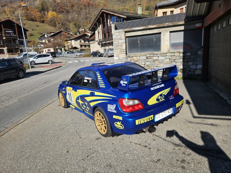 K&ouml;nnte beinhalten: Ein blaues Subaru Rallye-Auto mit gelben Akzenten und goldenen R&auml;dern steht auf einer Stra&szlig;e. Das Auto hat einen gro&szlig;en Heckspoiler und Sponsorenaufkleber. Im Hintergrund sind Geb&auml;ude und B&auml;ume zu sehen.