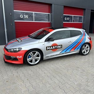 Puede incluir: Un Volkswagen Scirocco plateado con una decoración de Martini Racing. El coche tiene rayas azules, rojas y blancas en el lateral y un logotipo de Martini en la puerta. El coche está aparcado delante de una puerta de garaje roja con el número G 57.