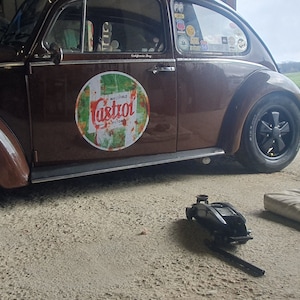 Puede incluir: Un Volkswagen Beetle vintage marrón con un logotipo de Castrol en el lateral. El coche está aparcado en un camino de grava con un gato y un cojín en primer plano.