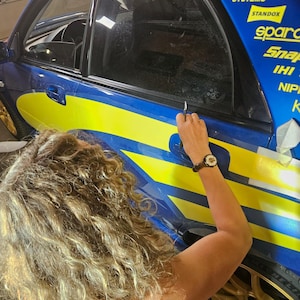 K&ouml;nnte beinhalten: Ein blau-gelbes Rennauto mit Sponsorenlogos wird bearbeitet. Eine Person verwendet ein Werkzeug, um an der Autoscheibe zu arbeiten. Das Auto hat goldene Felgen.