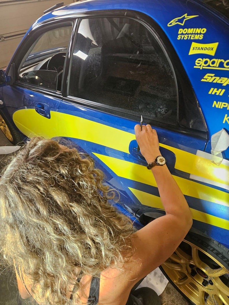 K&ouml;nnte beinhalten: Ein blau-gelbes Rennauto mit Sponsorenlogos wird bearbeitet. Eine Person verwendet ein Werkzeug, um an der Autoscheibe zu arbeiten. Das Auto hat goldene Felgen.