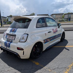 Peut inclure: Une petite voiture blanche avec des graphismes personnalisés et des roues noires est garée sur une surface pavée. La voiture a un aileron et une plaque d'immatriculation. La voiture a un design à rayures bleues et blanches sur le capot et le côté.
