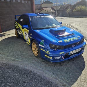 K&ouml;nnte beinhalten: Ein blaues Subaru Rallye-Auto mit goldenen Felgen und gelb-schwarzen Akzenten. Das Auto hat die Nummer 11 und verschiedene Sponsorenlogos. Auf der Front steht "Subaru World Rally Team".
