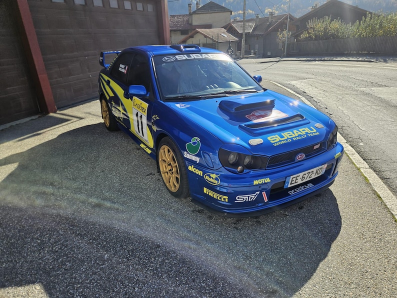 K&ouml;nnte beinhalten: Ein blaues Subaru Rallye-Auto mit goldenen Felgen und gelb-schwarzen Akzenten. Das Auto hat die Nummer 11 und verschiedene Sponsorenlogos. Auf der Front steht "Subaru World Rally Team".