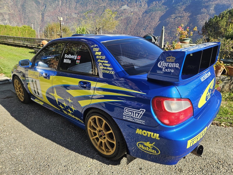 K&ouml;nnte beinhalten: Ein blaues Subaru Rallye-Auto mit gelben und wei&szlig;en Akzenten. Das Auto hat goldfarbene Felgen, Sponsorenaufkleber und einen gro&szlig;en Heckspoiler mit "Corona Extra"-Branding. Die Nummer "11" ist auf der Seite sichtbar.