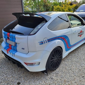 Peut inclure: Ford Focus RS blanche avec des jantes noires et un aileron arrière. La voiture présente des bandes de course bleues et rouges et la marque Martini sur le côté. Elle est garée sur un chemin de gravier.