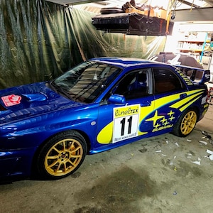 K&ouml;nnte beinhalten: Ein blaues Subaru-Rallyeauto mit goldenen Felgen und gelb-blauen Aufklebern. Das Auto hat die Nummer 11 auf der Seite und ein rot-wei&szlig;es Emblem auf der Motorhaube. Es steht in einer Garage.