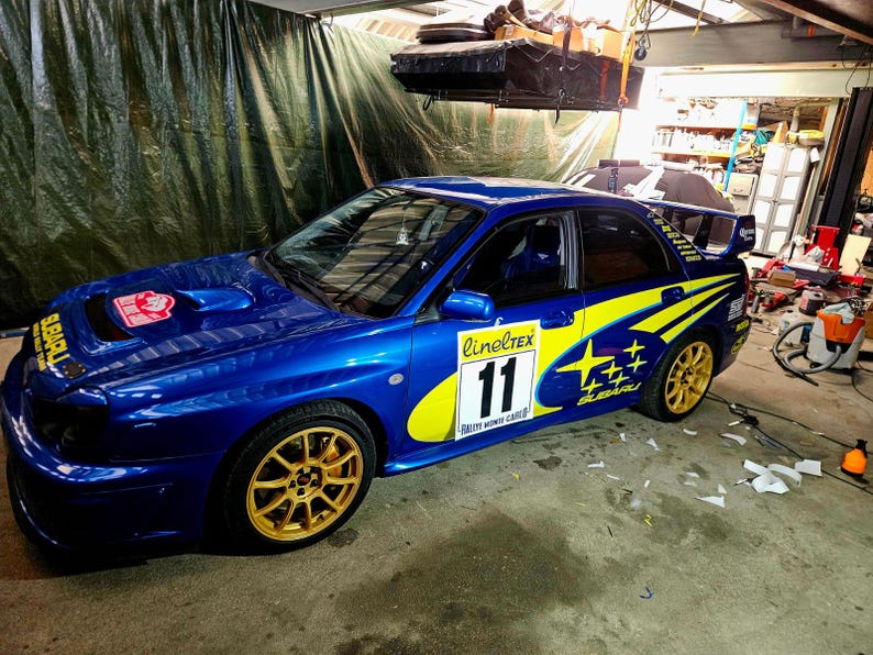 K&ouml;nnte beinhalten: Ein blaues Subaru-Rallyeauto mit goldenen Felgen und gelb-blauen Aufklebern. Das Auto hat die Nummer 11 auf der Seite und ein rot-wei&szlig;es Emblem auf der Motorhaube. Es steht in einer Garage.
