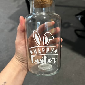 May include: Clear glass bottle with a cork lid and a white bunny ear design. The bottle has the text "Happy Easter" printed on it.