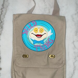 May include: A tan canvas messenger bag with a blue and pink embroidered patch featuring a cartoon frog with the text "Wild Thing".