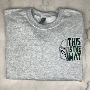 Op de afbeelding: Een opgevouwen lichtgrijs sweatshirt met een ronde hals. Het ontwerp op de voorkant heeft een helmomtrek en de tekst "THIS IS THE WAY" in groen en zwart.