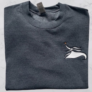 Może przedstawiać: Szary t-shirt z haftowanym białym obrazkiem Zero, postacią z filmu "Koszmar przed świętami". Zero trzyma w pysku świecącą pomarańczową kulę i leci w powietrzu.