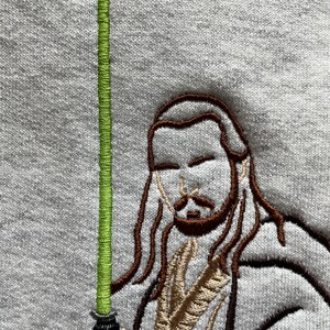 May include: Embroidered image of a Jedi Knight holding a green lightsaber. The Jedi Knight is wearing a brown robe and has long brown hair.