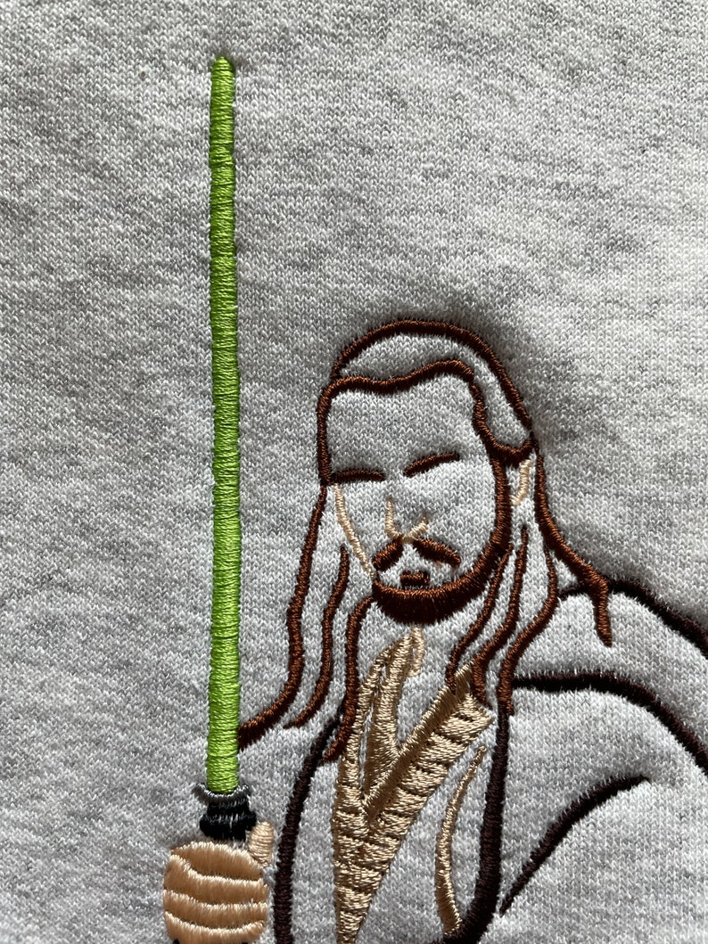 May include: Embroidered image of a Jedi Knight holding a green lightsaber. The Jedi Knight is wearing a brown robe and has long brown hair.