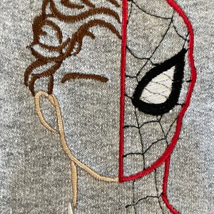 May include: Embroidered design on a gray sweatshirt featuring a split face of a person and a Spiderman mask. The person's hair is brown, and the Spiderman mask is red with black webbing and a white eye.