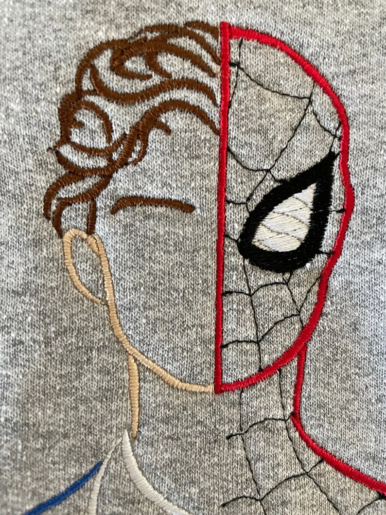 May include: Embroidered design on a gray sweatshirt featuring a split face of a person and a Spiderman mask. The person's hair is brown, and the Spiderman mask is red with black webbing and a white eye.