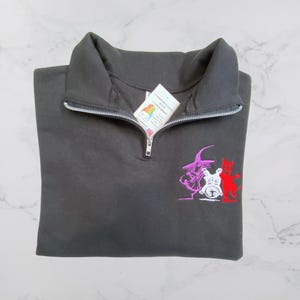 Może przedstawiać: Czarna bluza z zamkiem błyskawicznym na ćwierć długości z haftowanym wzorem przedstawiającym czarownicę, kota i ducha. Wzór jest w kolorze fioletowym, białym i czerwonym.