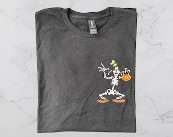 May include: A charcoal grey crew neck t-shirt featuring a graphic of a cartoon character dressed as a skeleton. The character is white with orange shoes and is holding an orange jack-o'-lantern bucket. The shirt has a Gildan brand label.