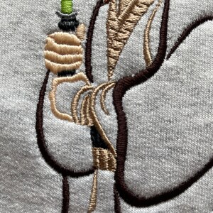 May include: Embroidered design of a person holding a lightsaber. The person is wearing a brown robe and has long brown hair. The lightsaber is green and black.