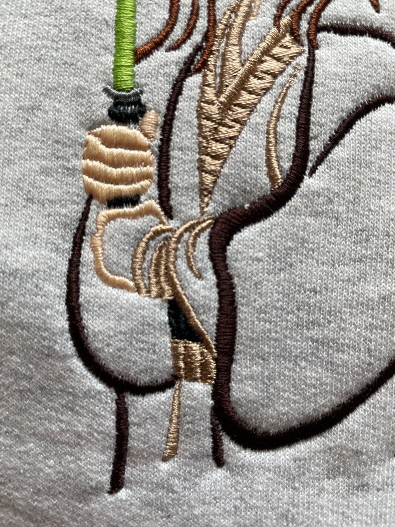May include: Embroidered design of a person holding a lightsaber. The person is wearing a brown robe and has long brown hair. The lightsaber is green and black.