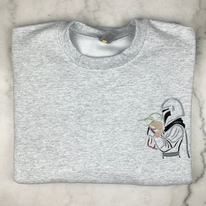 May include: A gray sweatshirt with an embroidered image of a character from the Star Wars franchise, The Mandalorian, holding a small creature, Grogu, also known as Baby Yoda.