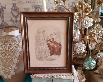 Könnte beinhalten: Ein gerahmter Vintage-Druck, der drei Frauen in historischer Kleidung zeigt, ruhend auf einer dekorativen Schachtel mit blau-weißem Toile-Muster. Die Schachtel steht auf einem hellblauen, verzierten Tisch. Ein Paar cremefarbene Schuhe mit dekorativen Schnallen ist ebenfalls zu sehen.