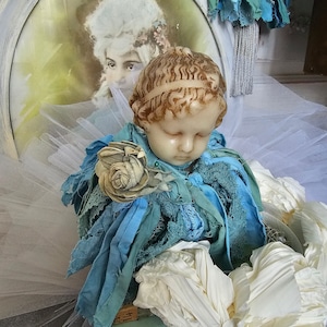 Könnte beinhalten: Eine Vintage-Skulptur eines cherubischen Engels mit geschlossenen Augen, geschmückt mit blauem Stoff und einer cremefarbenen Rose. Die Skulptur ruht auf einem Stapel antiker Bücher, mit einem gerahmten Porträt im Hintergrund. Die Farbpalette umfasst Blau-, Creme- und Brauntöne.