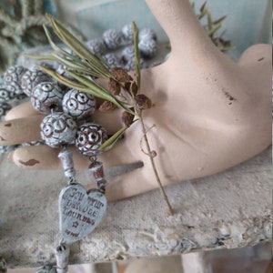 May include: A rosary with white and brown beads, held by a cream-colored hand statue. The rosary has a heart-shaped pendant with text. A sprig of greenery rests in the hand. The background is a light blue and white.
