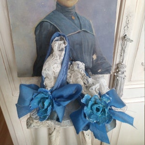 Könnte beinhalten: Ein antikes Porträt einer Frau in einem blauen Kleid bildet den Hintergrund für ein dekoratives Element. Das Element umfasst eine blaue Schleife und eine blaue Papierblume mit weißen Spitzendetails. Der Stil erinnert an eine vergangene Ära.