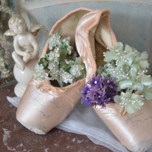 May include: A pair of vintage ballet pointe shoes in a soft pink hue, filled with delicate white and purple floral arrangements. The shoes have handwritten inscriptions and rest on a marble surface, evoking a sense of nostalgia and grace.