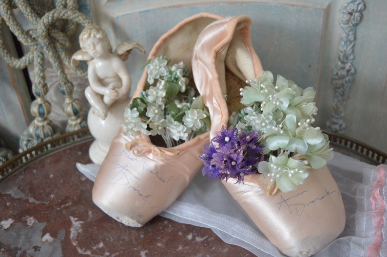 May include: A pair of vintage ballet pointe shoes in a soft pink hue, filled with delicate white and purple floral arrangements. The shoes have handwritten inscriptions and rest on a marble surface, evoking a sense of nostalgia and grace.