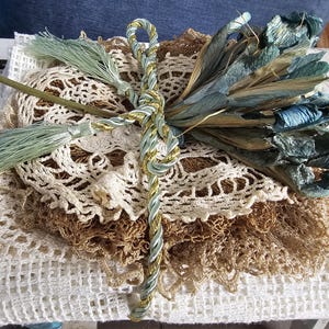 Könnte beinhalten: Eine Sammlung von Vintage-Spitzendeckchen in Weiß- und Beigetönen, gekrönt von einem Bund getrockneter blauer Blumen und einem dekorativen Kordel. Die Anordnung ist mit einer gedrehten Kordel in Gold, Blau und Weiß gebunden.