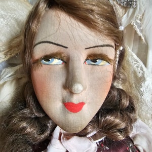 May include: Close-up of a vintage doll's face. The doll has painted features, including blue eyes, red lips, and blushed cheeks. Her brown hair is styled in curls. The doll is wearing a burgundy dress with a white collar.
