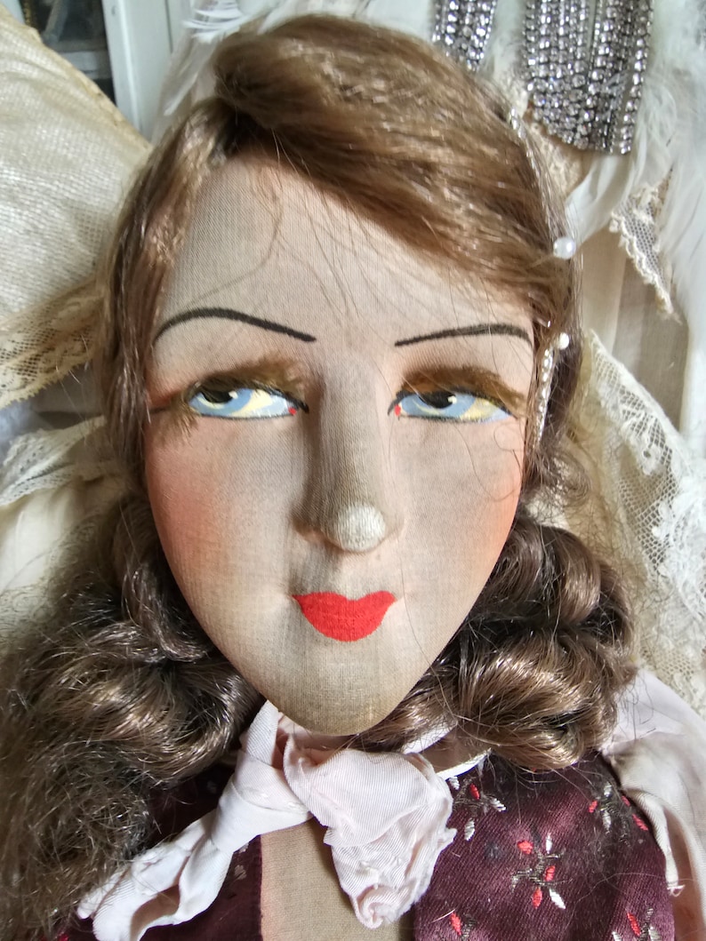 May include: Close-up of a vintage doll's face. The doll has painted features, including blue eyes, red lips, and blushed cheeks. Her brown hair is styled in curls. The doll is wearing a burgundy dress with a white collar.