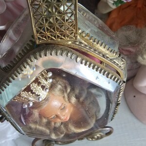 May include: A vintage, hexagonal-shaped trinket box with a clear glass exterior and ornate gold-colored metal trim. Inside, a doll's head with blonde hair and a crown is visible. The box has a hinged lid and decorative filigree details.