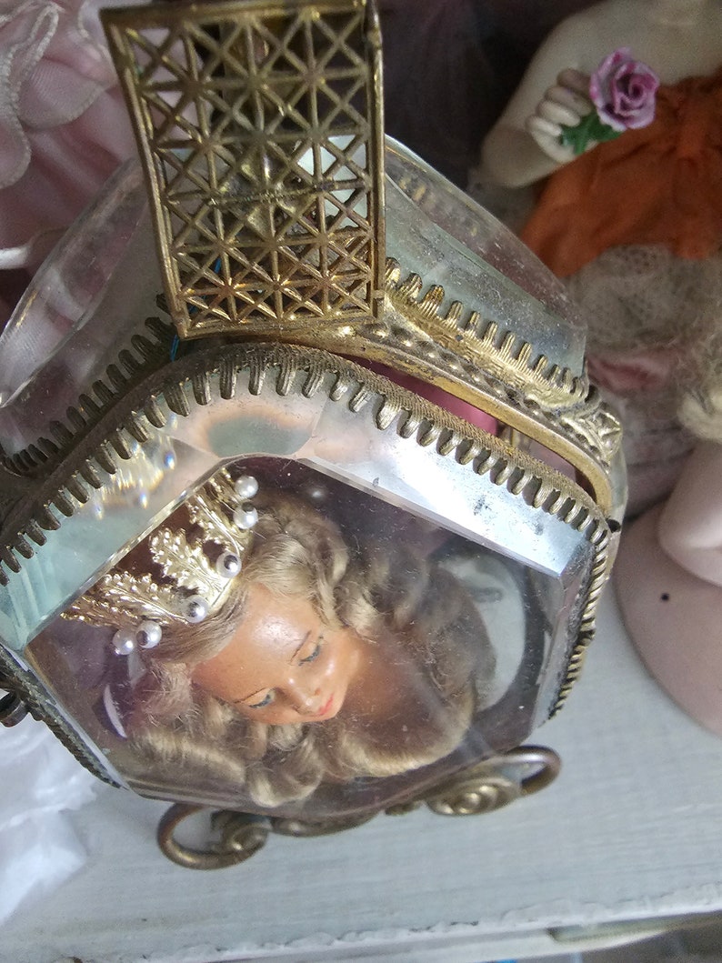 May include: A vintage, hexagonal-shaped trinket box with a clear glass exterior and ornate gold-colored metal trim. Inside, a doll's head with blonde hair and a crown is visible. The box has a hinged lid and decorative filigree details.